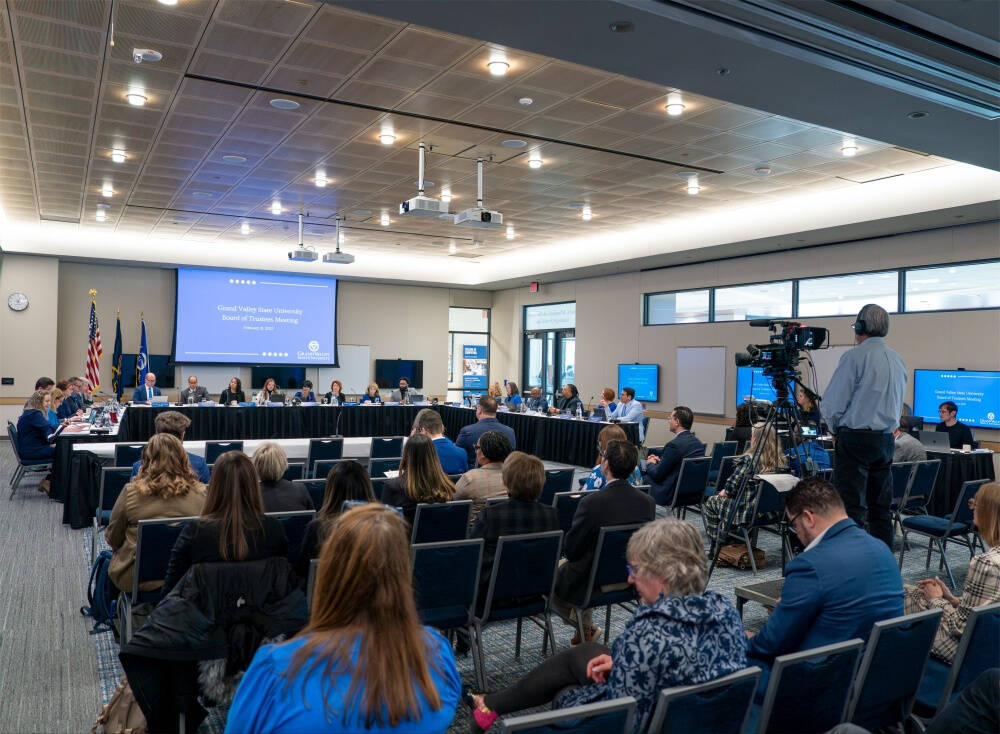 A wide-angle view of a group of people in a meeting held in a large, modern conference room. Some are seated along U-shaped table at the front, facing the rest. A large projection screen displays in the background.